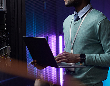 A man holding a laptop in a server room, surrounded by tech equipment