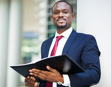 A smiling black male, dressed in professional attaire, stands outside a building and holding an open notebook