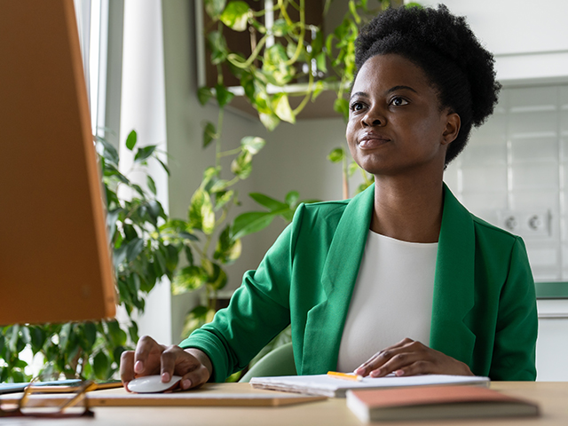 Focused African American woman working sits at computer desk holding mouse in hand and looking at monitor