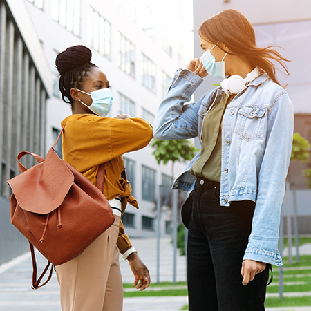 Young cheerful female friends in masks meeting outside and greeting with elbow touch