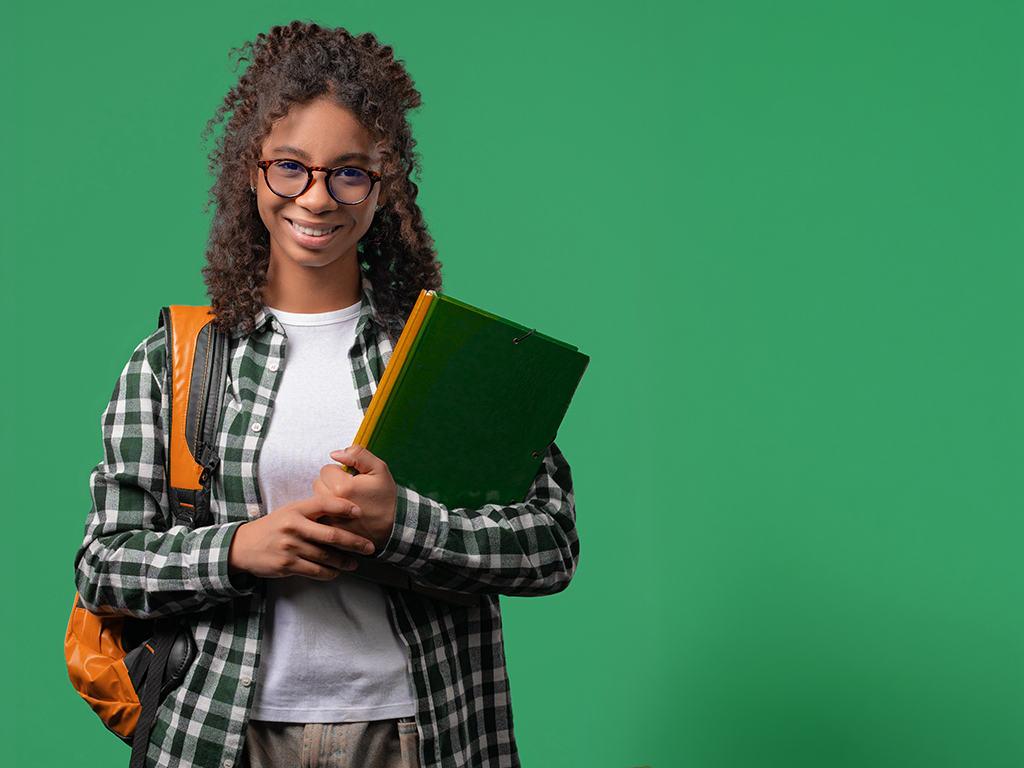 Smiling young African American female student wearing an orange shirt and white t-shirt with blue backpack on her shoulder and an orange book in her hand on a green background.