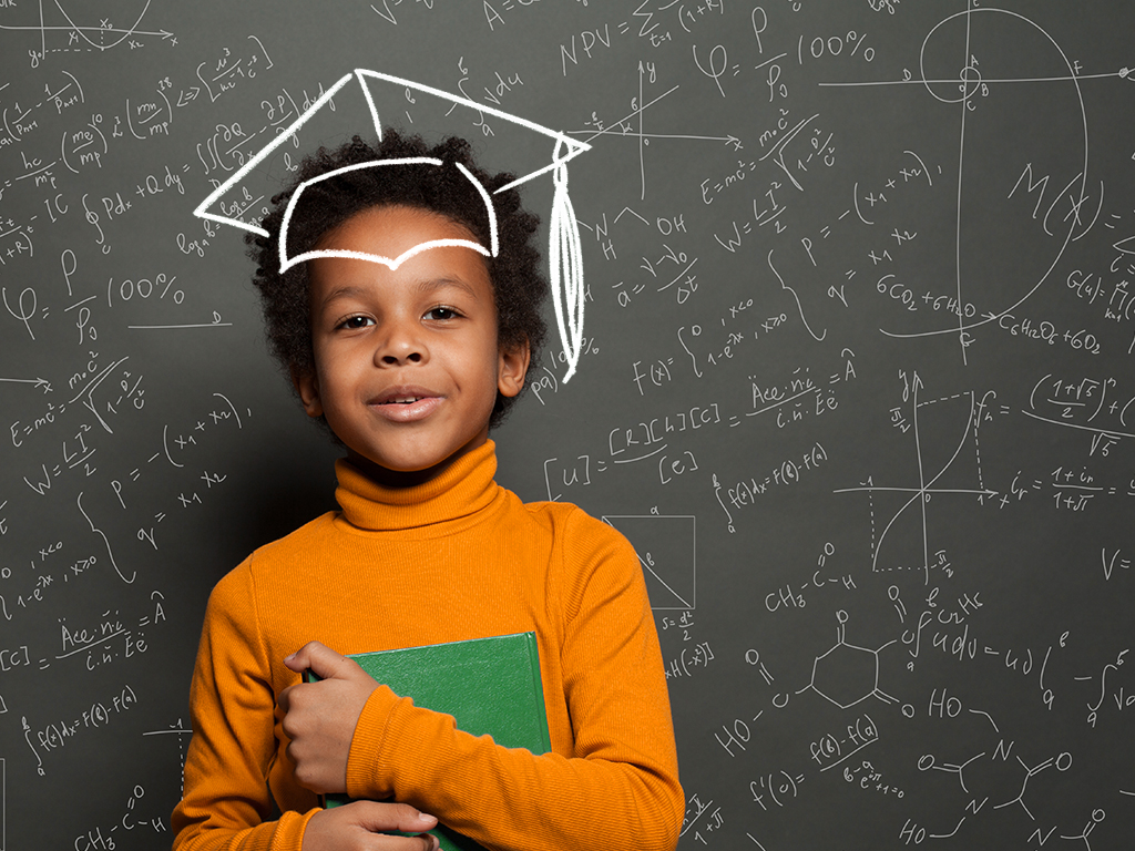 African American child holding a book, standing in front of a black chalkboard with science and maths formulas. A chalk drawing of a graduation cap is placed over the childs head.